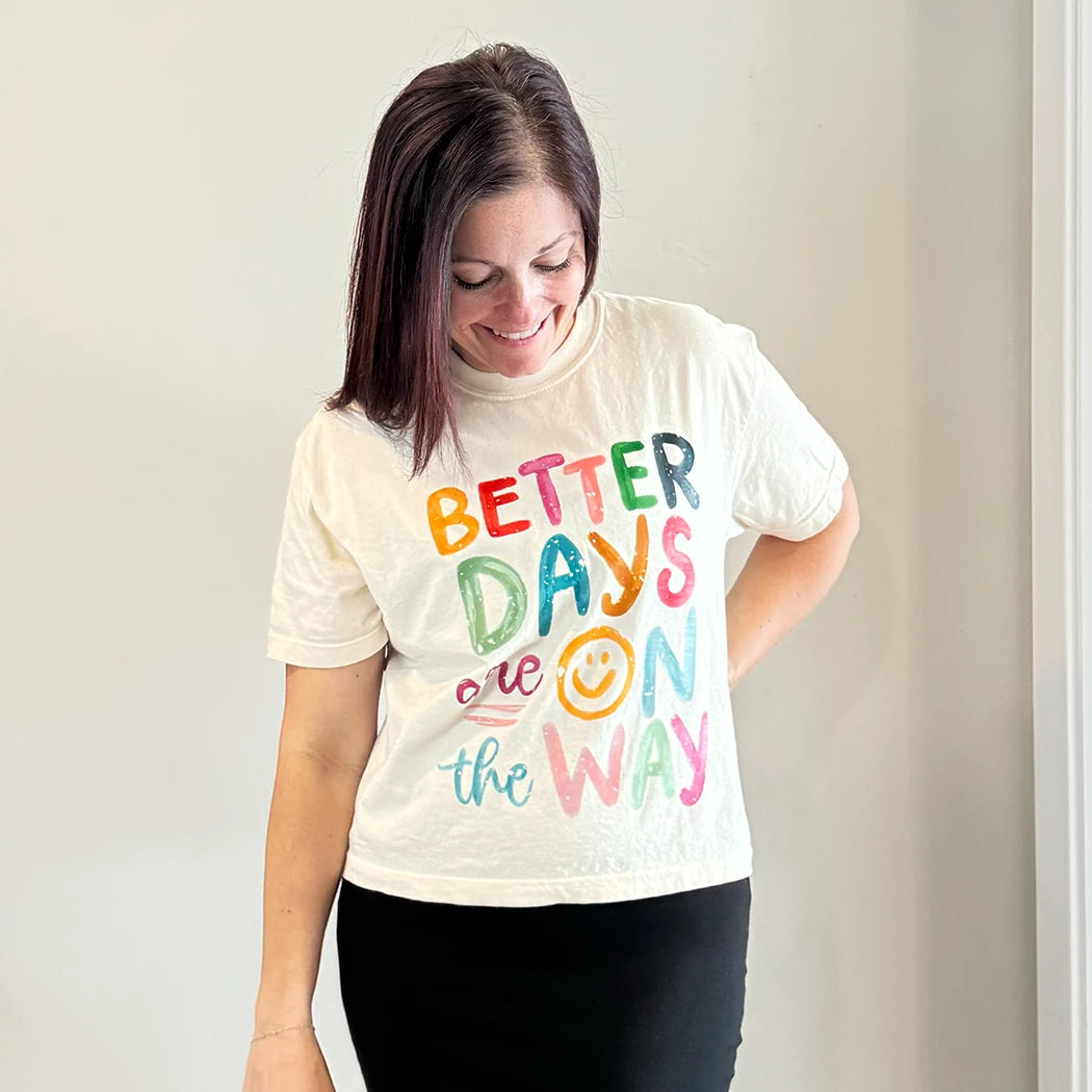 A light-skinned person with shoulder-length dark hair smiles, wearing a womens graphic tshirts, cream-colored t-shirt with the text "BETTER DAYS are ON the WAY" stacked in colorful, slightly uneven letters. A yellow smiley face replaces the 'O' in 'ON'.