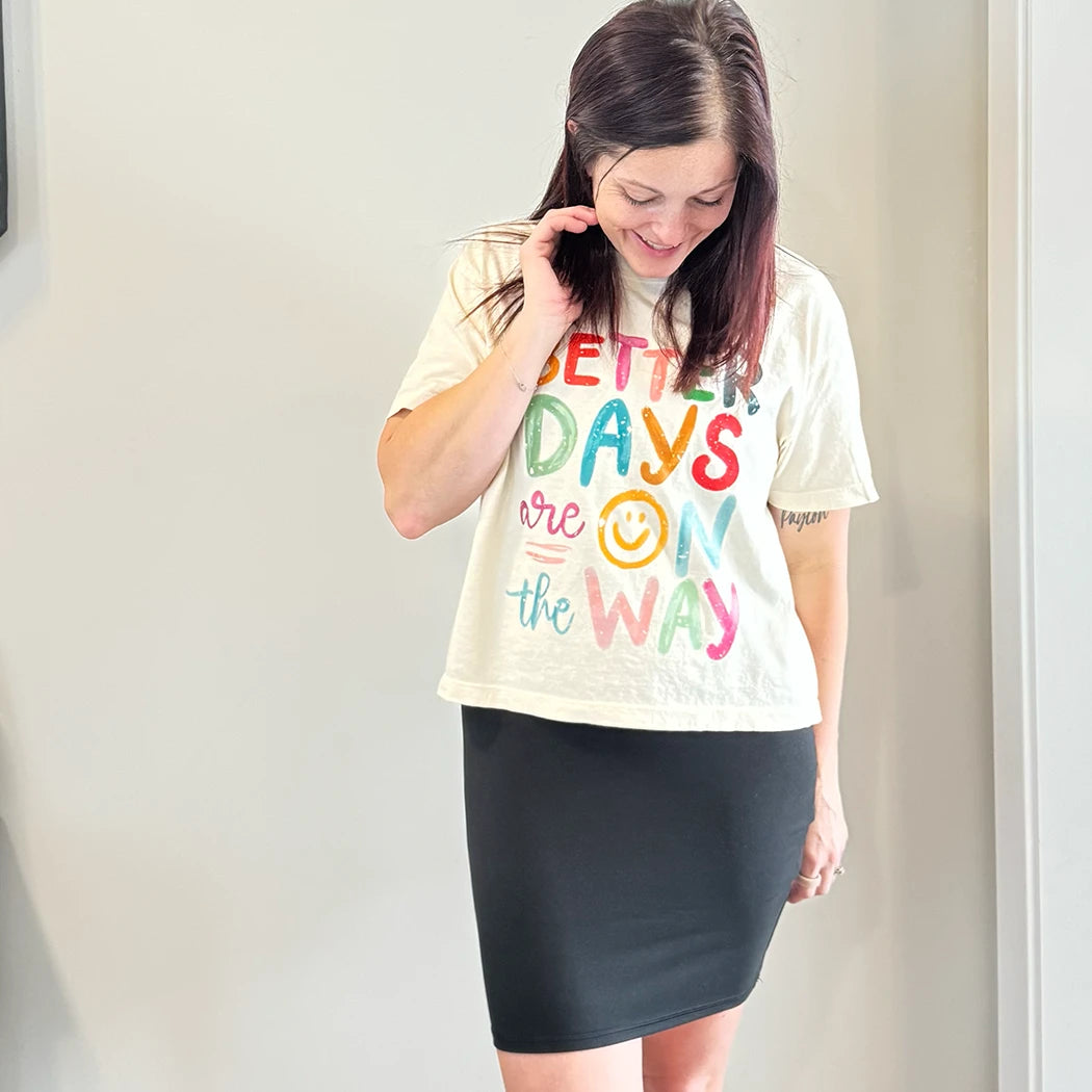 A light-skinned person with shoulder-length dark hair smiles, wearing a , womens graphic tshirtscream-colored t-shirt with the text "BETTER DAYS are ON the WAY" stacked in colorful, slightly uneven letters. A yellow smiley face replaces the 'O' in 'ON'.