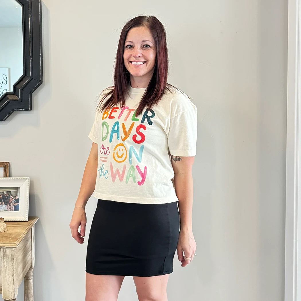 A light-skinned person with shoulder-length dark hair smiles, wearing a womens graphic tshirts, cream-colored t-shirt with the text "BETTER DAYS are ON the WAY" stacked in colorful, slightly uneven letters. A yellow smiley face replaces the 'O' in 'ON'.