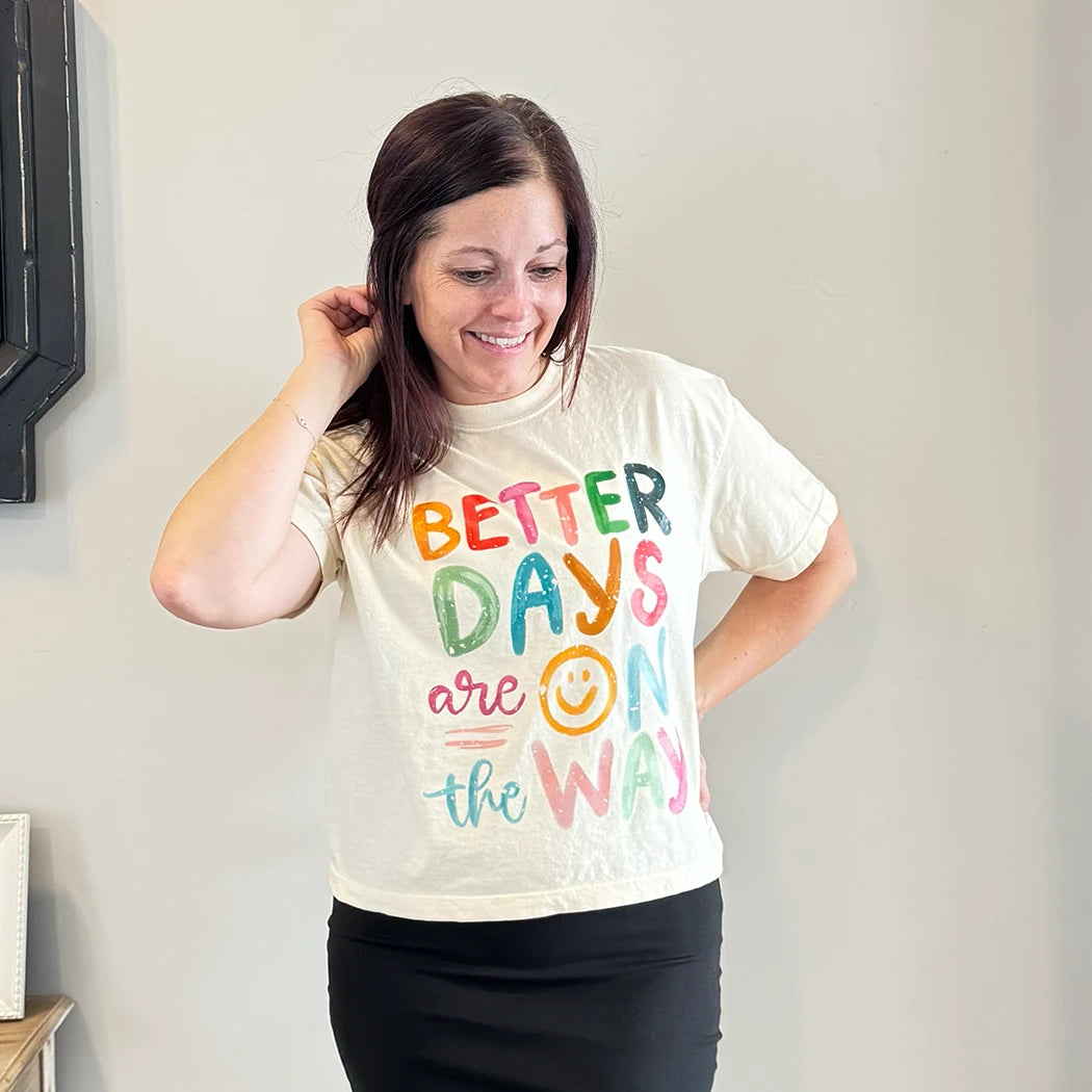 A light-skinned person with shoulder-length dark hair smiles, wearing a womens graphic tshirts, cream-colored t-shirt with the text "BETTER DAYS are ON the WAY" stacked in colorful, slightly uneven letters. A yellow smiley face replaces the 'O' in 'ON'.