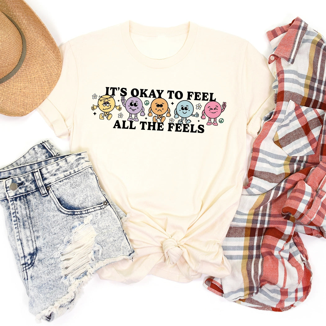 It’s okay to feel all the feels positive message shirt with smiley face characters. White shirt with colorful design. Flowers, peace sign, green, yellow, blue and orange design.
