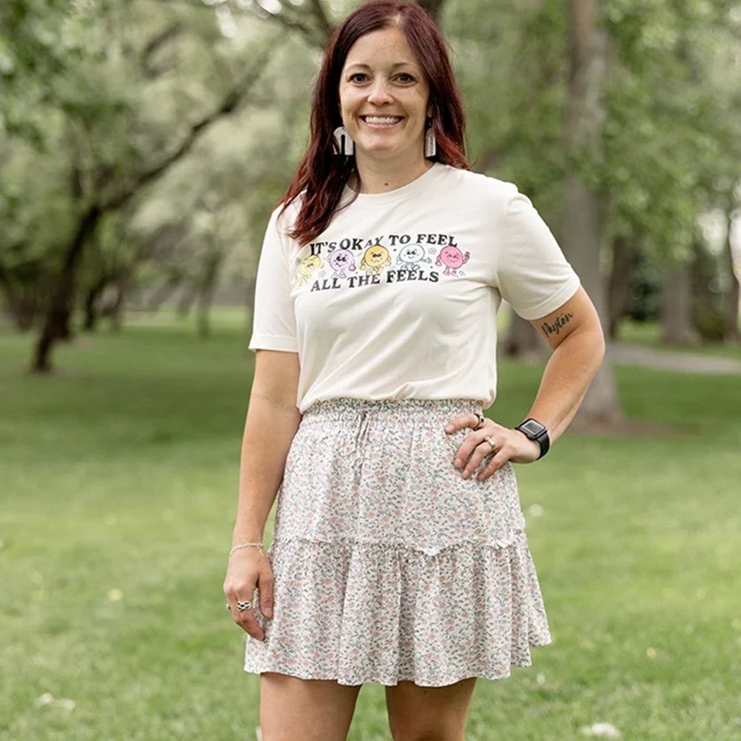It’s okay to feel all the feels positive message shirt with smiley face characters. White shirt with colorful design. Flowers, peace sign, green, yellow, blue and orange design.