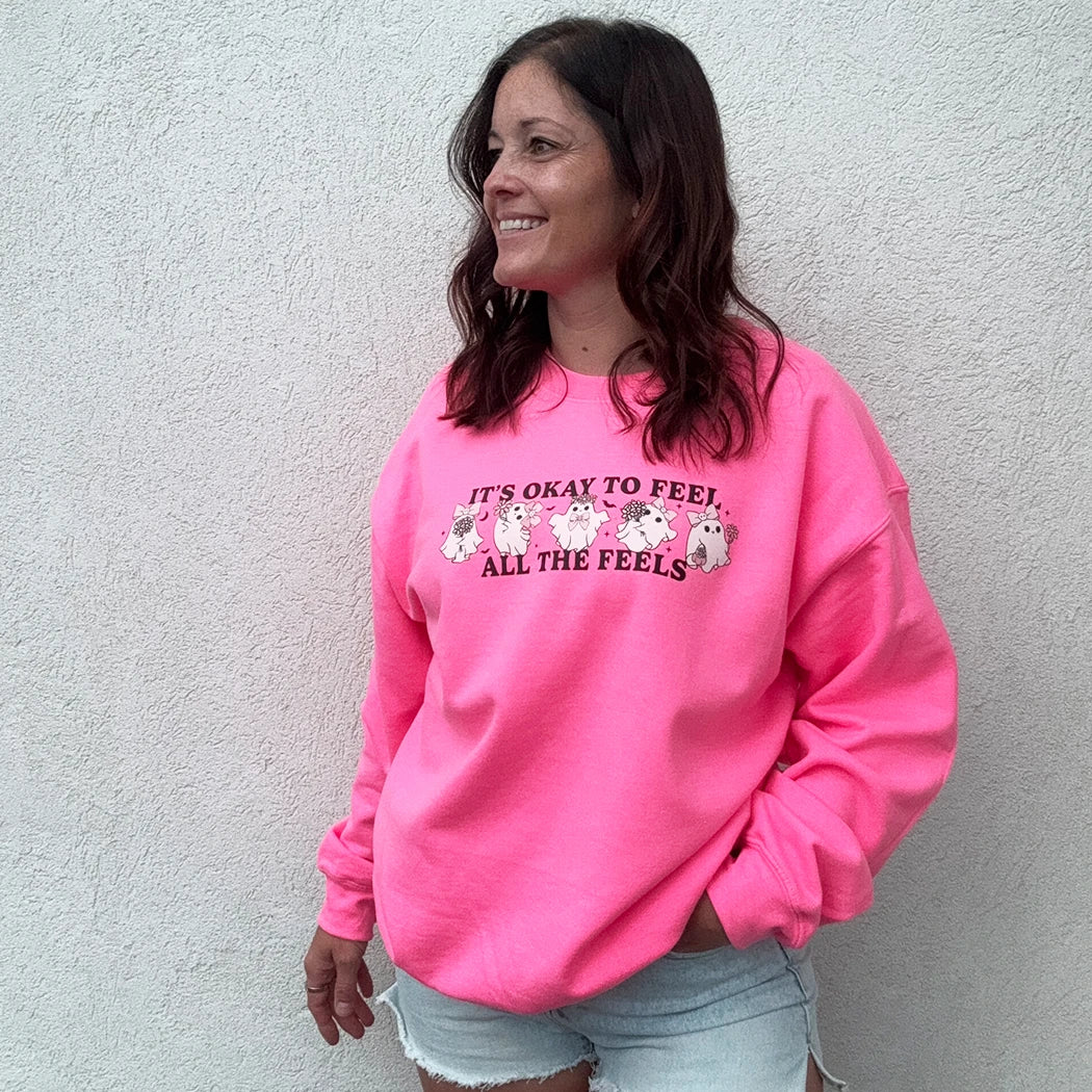 A woman stands against a light-colored wall, smiling and looking to the side. She is wearing a bright, neon pink crewneck with the phrase "IT'S OKAY TO FEEL ALL THE FEELS" and a line of small, multi-colored characters across the chest. She has her hand in the pocket of her light blue denim shorts.