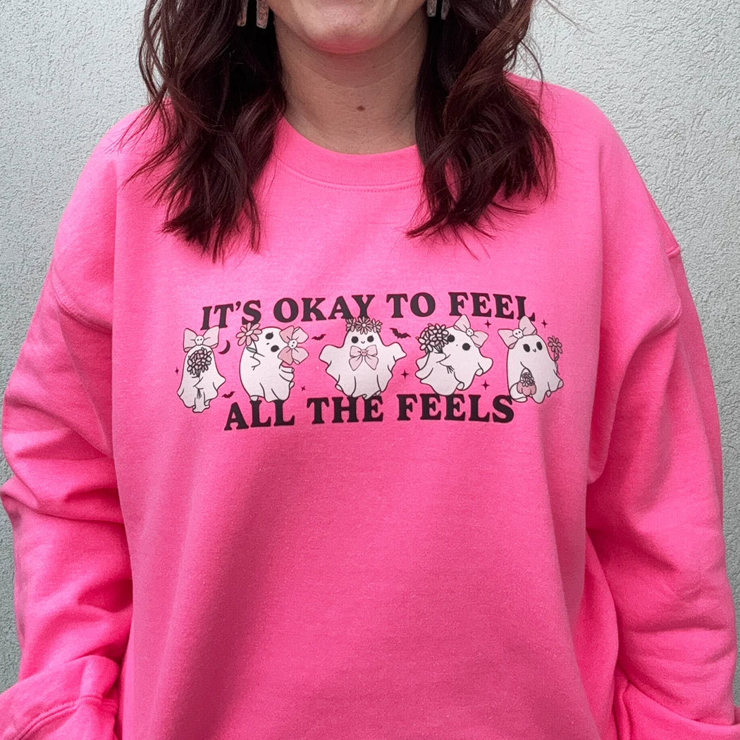 A woman stands against a light-colored wall, smiling and looking to the side. She is wearing a bright, neon pink crewneck with the phrase "IT'S OKAY TO FEEL ALL THE FEELS" and a line of small, multi-colored characters across the chest. She has her hand in the pocket of her light blue denim shorts.