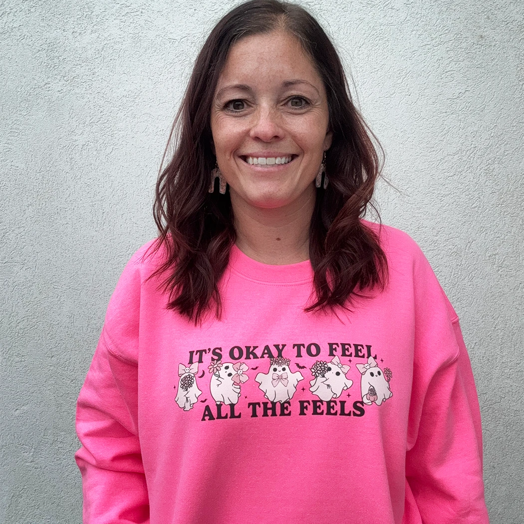 A woman stands against a light-colored wall, smiling and looking to the side. She is wearing a bright, neon pink crewneck with the phrase "IT'S OKAY TO FEEL ALL THE FEELS" and a line of small, multi-colored characters across the chest. She has her hand in the pocket of her light blue denim shorts.
