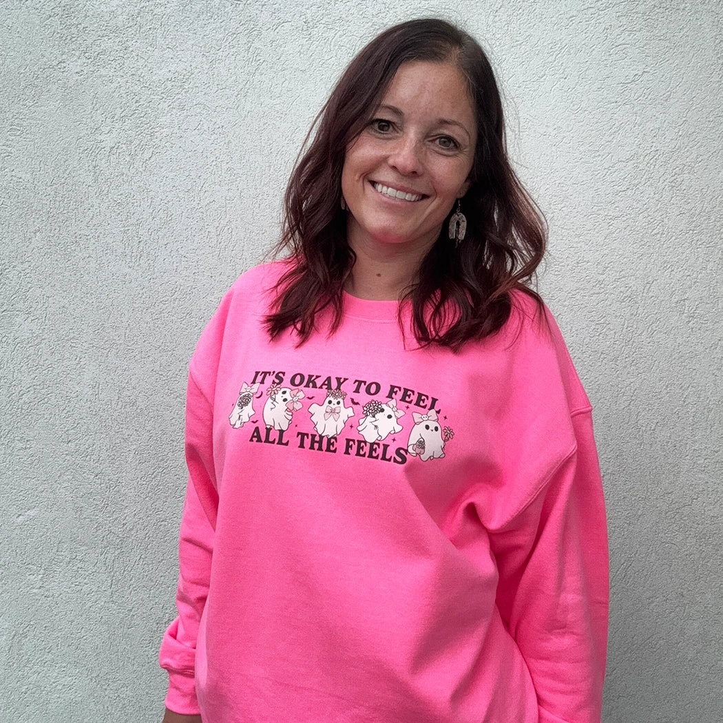 A woman stands against a light-colored wall, smiling and looking to the side. She is wearing a bright, neon pink crewneck with the phrase "IT'S OKAY TO FEEL ALL THE FEELS" and a line of small, multi-colored characters across the chest. She has her hand in the pocket of her light blue denim shorts.