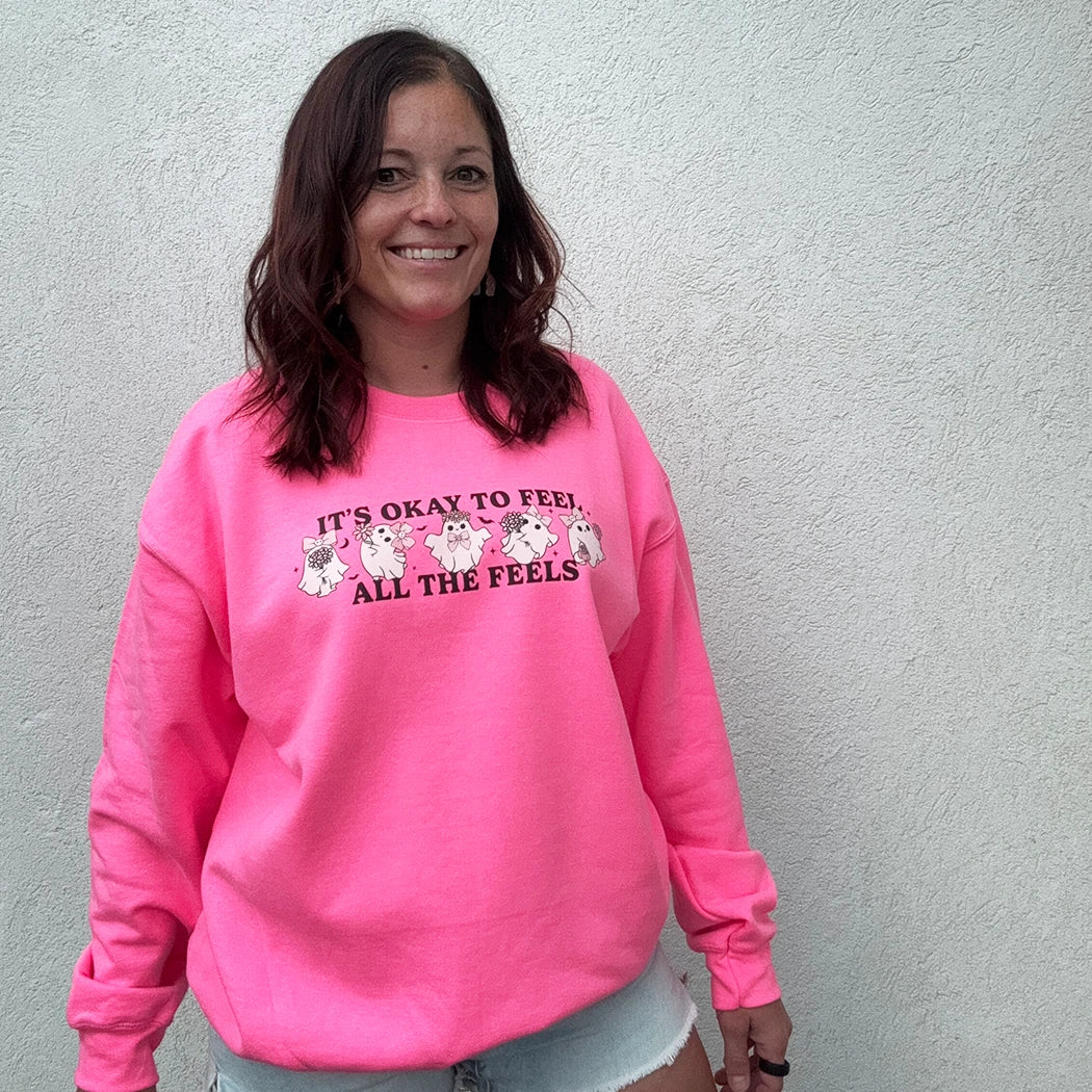 A woman stands against a light-colored wall, smiling and looking to the side. She is wearing a bright, neon pink crewneck with the phrase "IT'S OKAY TO FEEL ALL THE FEELS" and a line of small, multi-colored characters across the chest. She has her hand in the pocket of her light blue denim shorts.