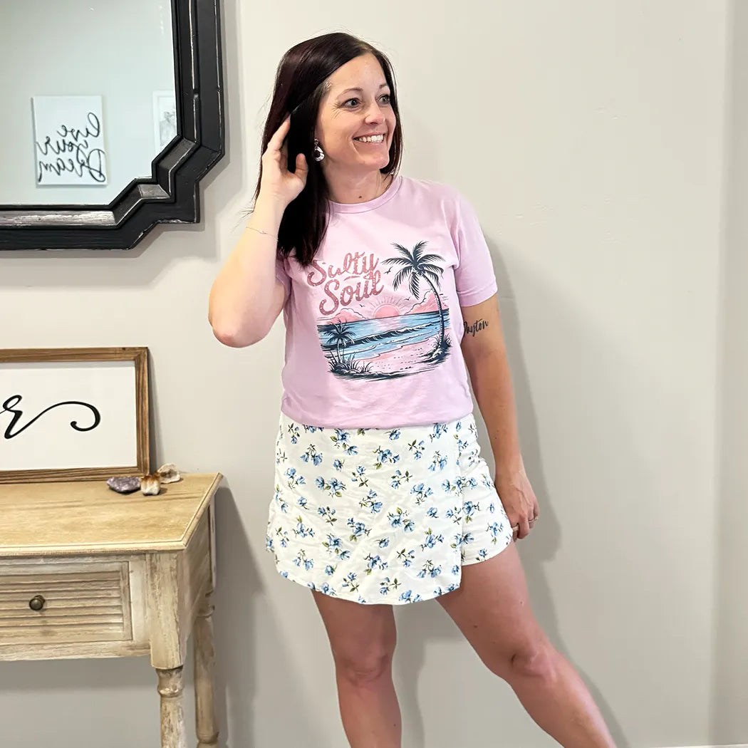 A fair-skinned person with shoulder-length dark hair smiles, wearing a light purple graphic apparel t-shirt with a beach-themed graphic. The graphic includes a sunset with pink and orange hues over a blue ocean, a silhouetted palm tree, and the cursive text "Salty Soul" arched above the scene.