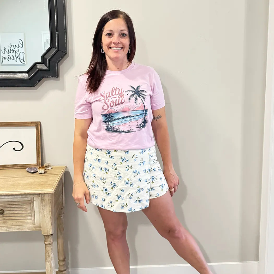 A fair-skinned person with shoulder-length dark hair smiles, wearing a light purple graphic apparel t-shirt with a beach-themed graphic. The graphic includes a sunset with pink and orange hues over a blue ocean, a silhouetted palm tree, and the cursive text "Salty Soul" arched above the scene.