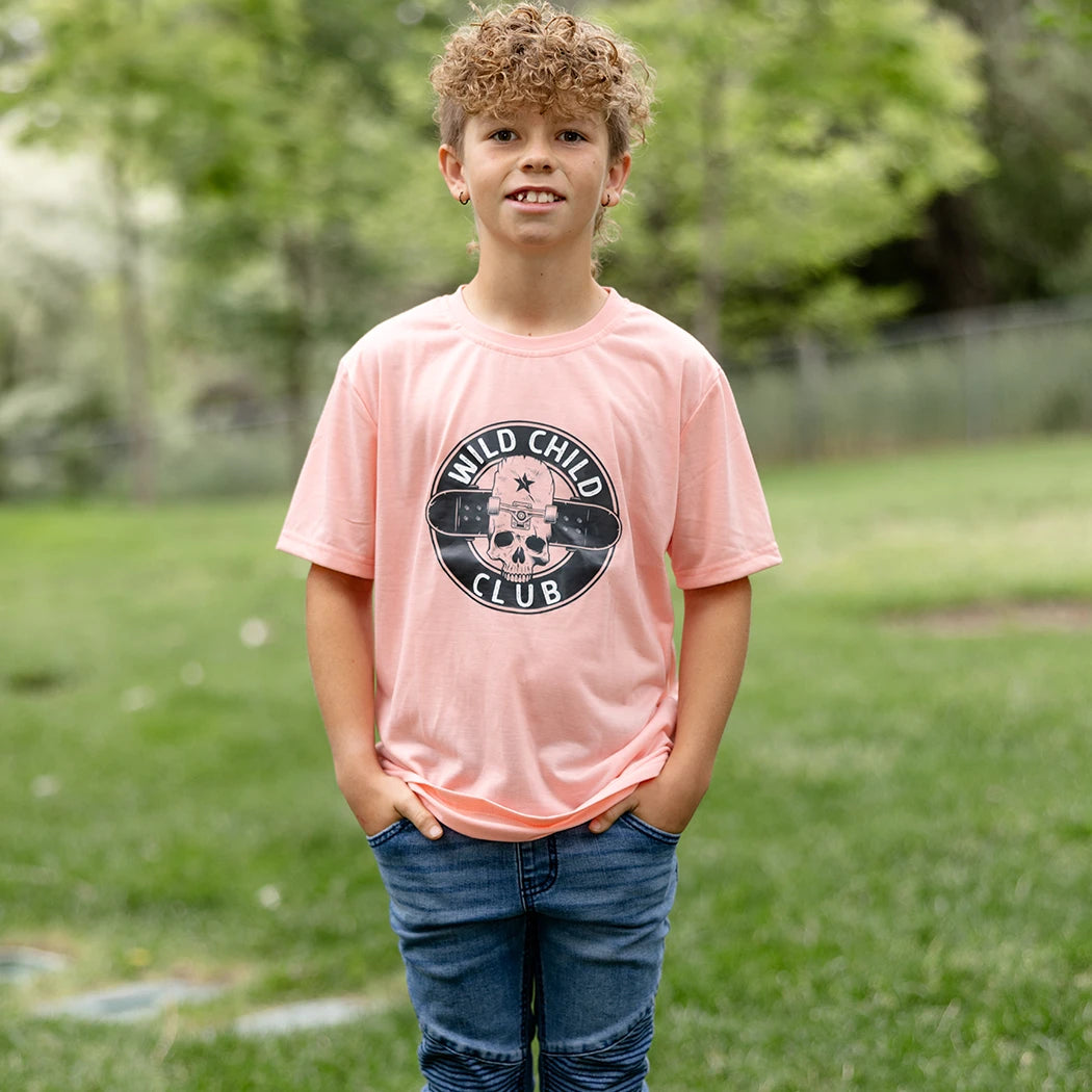 A young boy with blonde curly hair and smile stands with his hands in his pockets, wearing a light peach short-sleeved graphic apparel t-shirt with a circular black and white 'Wild Child Club' graphic. The graphic includes a skull with a star, wings, and the text 'WILD CHILD CLUB'. He is also wearing blue denim jeans with distressed details and is standing on a green lawn with blurred trees in the background.