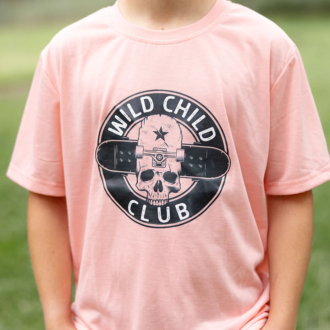A young boy with blonde curly hair and smile stands with his hands in his pockets, wearing a light peach short-sleeved graphic apparel t-shirt with a circular black and white 'Wild Child Club' graphic. The graphic includes a skull with a star, wings, and the text 'WILD CHILD CLUB'. He is also wearing blue denim jeans with distressed details and is standing on a green lawn with blurred trees in the background.