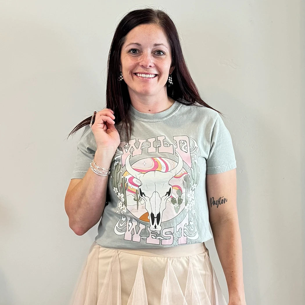 A fair-skinned person with shoulder-length dark hair smiles, wearing a womens graphic tshirts, light green graphic apparel t-shirt with a stylized graphic in muted pinks, oranges, and greens. The graphic includes a cow skull with desert elements like cacti and a wavy landscape behind it, and the text "WILD" above and "WEST" below the central image. The design is western style.
