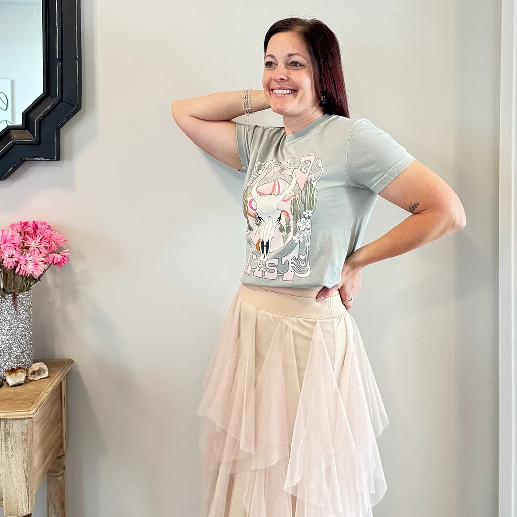 A fair-skinned person with shoulder-length dark hair smiles, wearing a womens graphic tshirts, light green graphic apparel t-shirt with a stylized graphic in muted pinks, oranges, and greens. The graphic includes a cow skull with desert elements like cacti and a wavy landscape behind it, and the text "WILD" above and "WEST" below the central image. The design is western style.