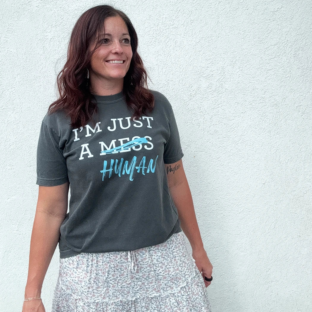 A person stands in front of a white wall, wearing a dark gray t-shirt with a white and blue graphic. The text reads "I'M JUST A MESS HUMAN," with the word "mess" having a blue line drawn through it and "human" written in blue cursive underneath.