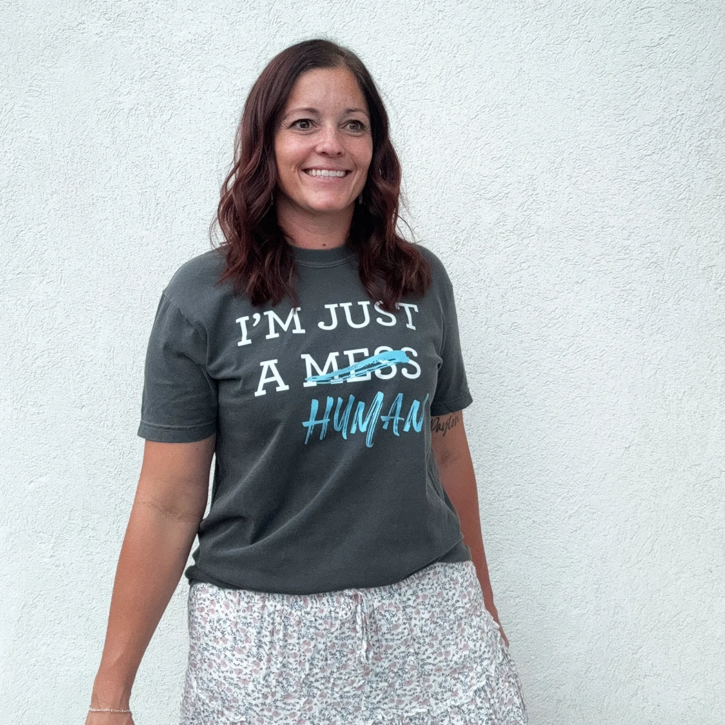A person stands in front of a white wall, wearing a dark gray t-shirt with a white and blue graphic. The text reads "I'M JUST A MESS HUMAN," with the word "mess" having a blue line drawn through it and "human" written in blue cursive underneath.