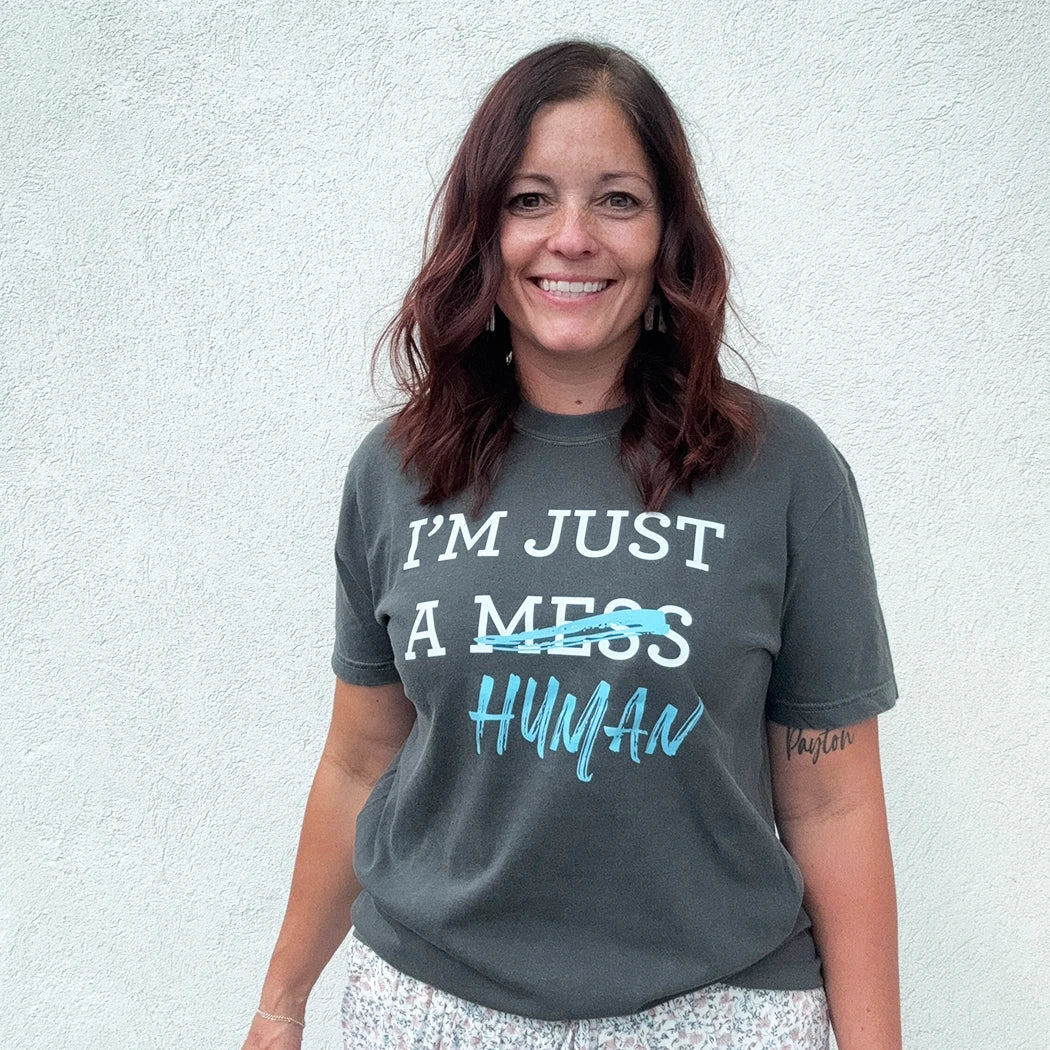 A person stands in front of a white wall, wearing a dark gray t-shirt with a white and blue graphic. The text reads "I'M JUST A MESS HUMAN," with the word "mess" having a blue line drawn through it and "human" written in blue cursive underneath.