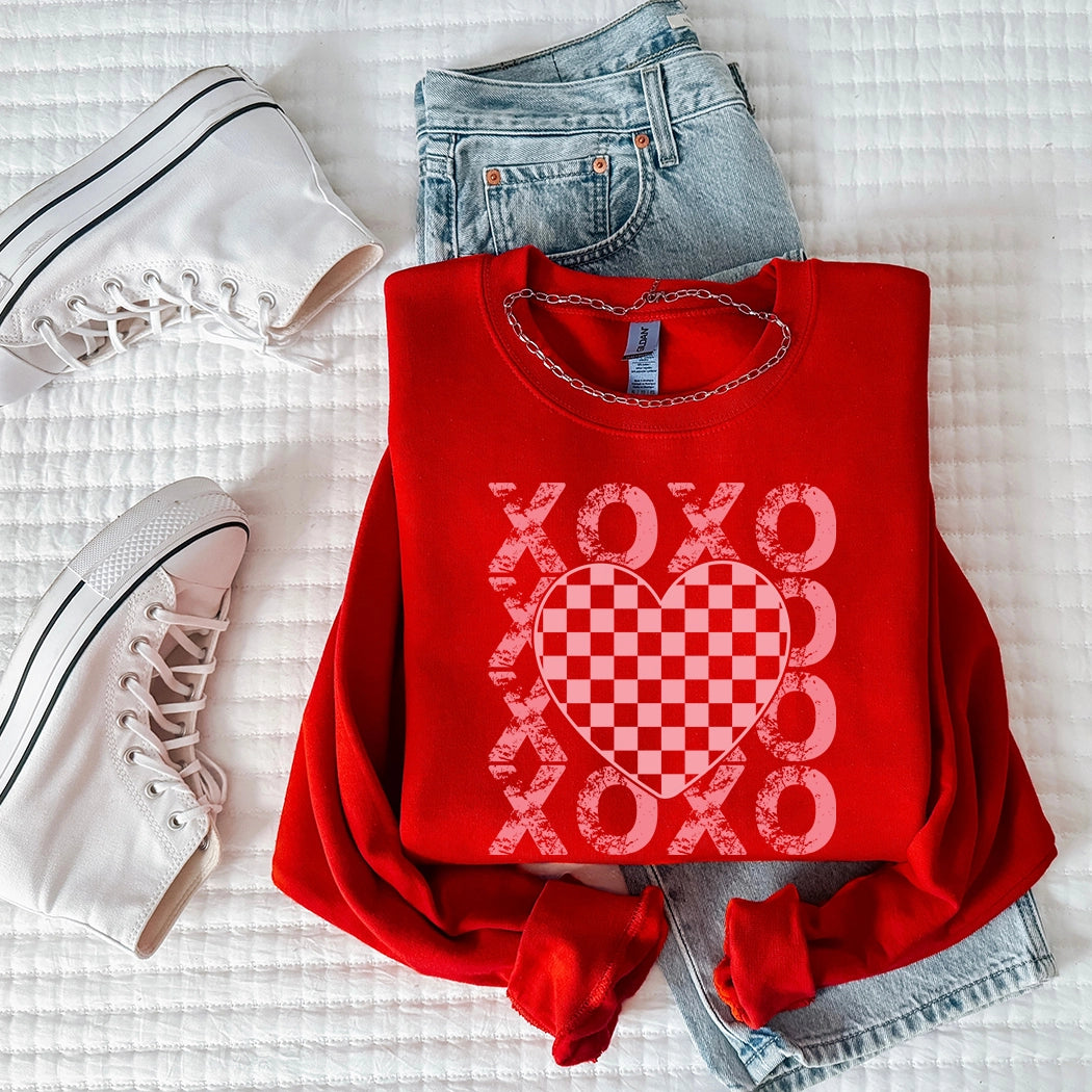 Flat lay of a bright red graphic apparel crewneck sweatshirt featuring a pink and white checkered heart graphic with distressed white 'XOXO' text vertically stacked on either side. The sweatshirt is styled with light wash denim jeans and a pair of white high-top sneakers on a white quilted blanket.