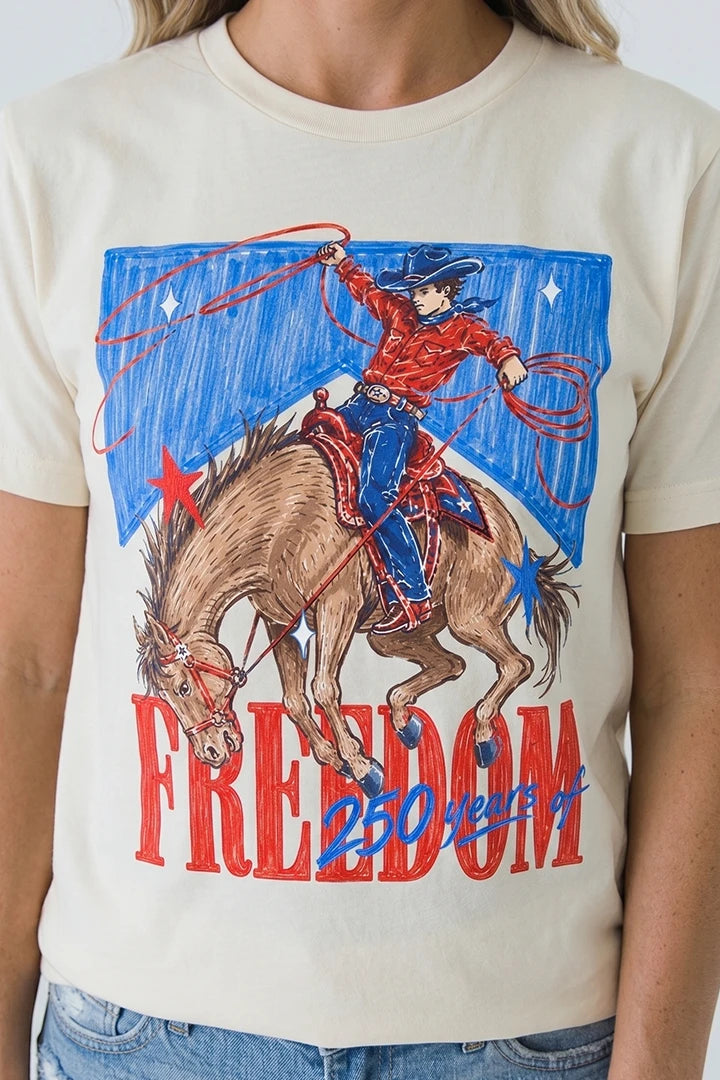 Close-up of a person wearing a cream-colored short-sleeved t-shirt with a vintage-style sketch graphic. The design depicts a cowboy in a red shirt and blue hat riding a brown bucking horse with a lasso, set against a blue block background with white stars. At the bottom, 'FREEDOM' is written in large red letters with '250 years of' overlaid in blue script.