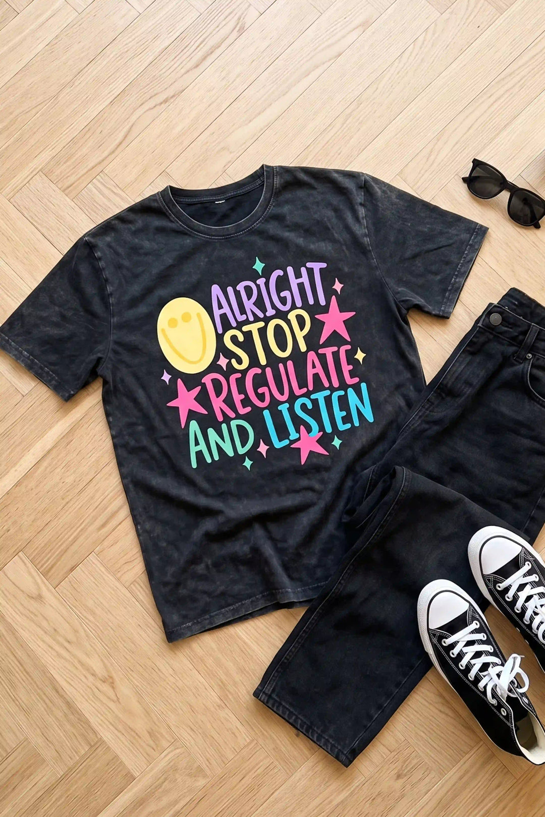 Dark washed black graphic t-shirt laid flat featuring colorful text reading "ALRIGHT STOP REGULATE AND LISTEN" accented with pink stars, green sparkles, and a yellow smiley face