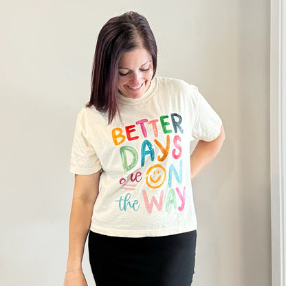 A light-skinned person with shoulder-length dark hair smiles, wearing a womens graphic tshirts, cream-colored t-shirt with the text "BETTER DAYS are ON the WAY" stacked in colorful, slightly uneven letters. A yellow smiley face replaces the 'O' in 'ON'.