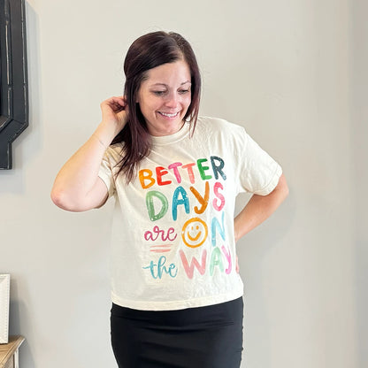 A light-skinned person with shoulder-length dark hair smiles, wearing a womens graphic tshirts, cream-colored t-shirt with the text "BETTER DAYS are ON the WAY" stacked in colorful, slightly uneven letters. A yellow smiley face replaces the 'O' in 'ON'.