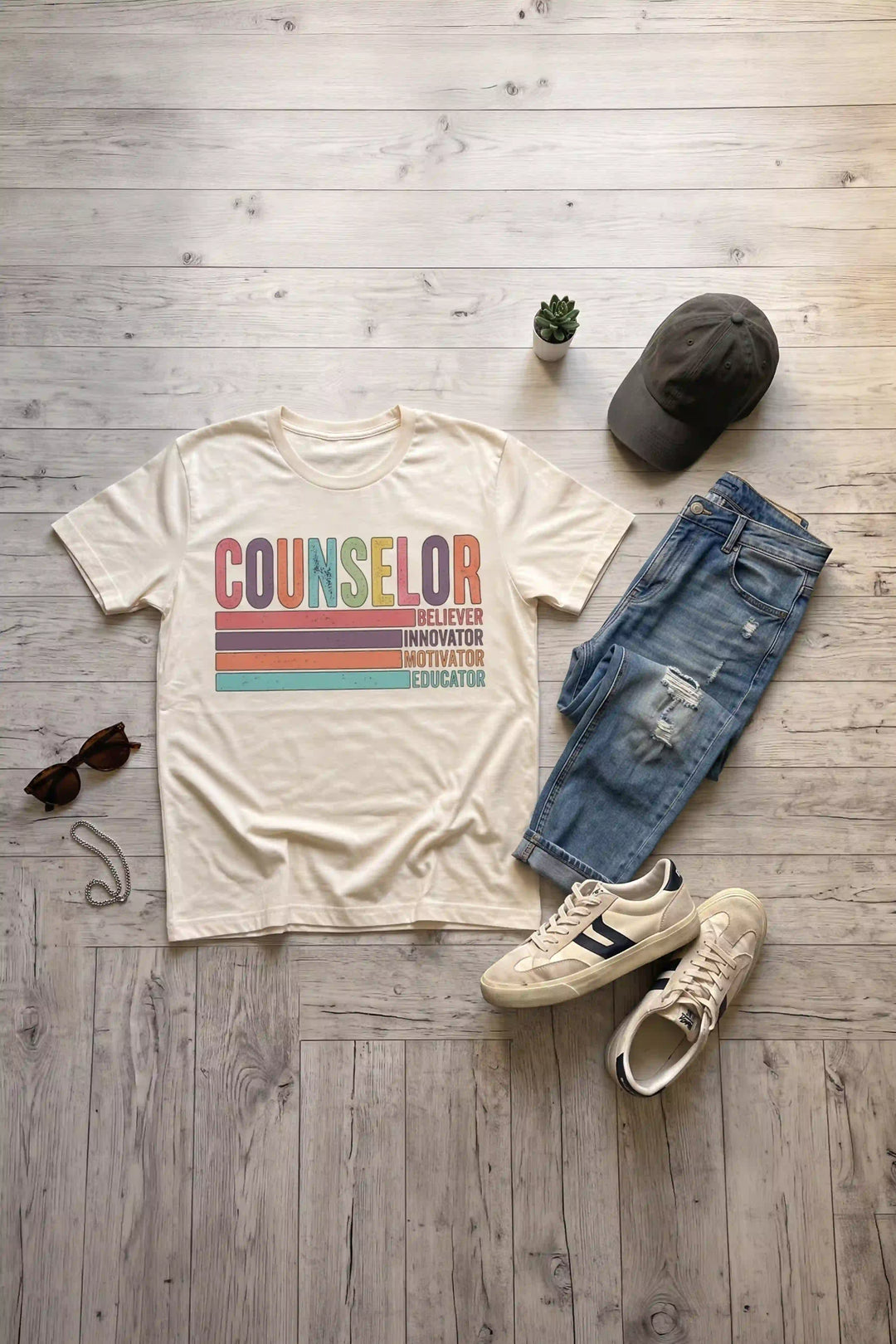 Cream retro graphic t-shirt laid flat featuring distressed colorful text reading "COUNSELOR" above color-blocked stripes labeled "BELIEVER, INNOVATOR, MOTIVATOR, EDUCATOR"