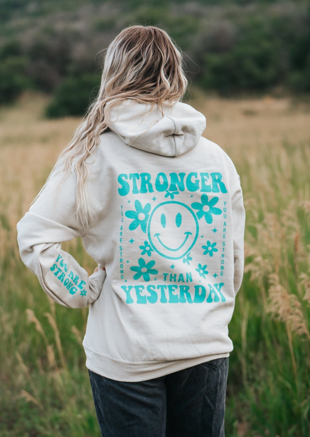 Model wearing sand color hoodie that says stronger than yesterday in a teal color. Design has flowers with smiley face. 