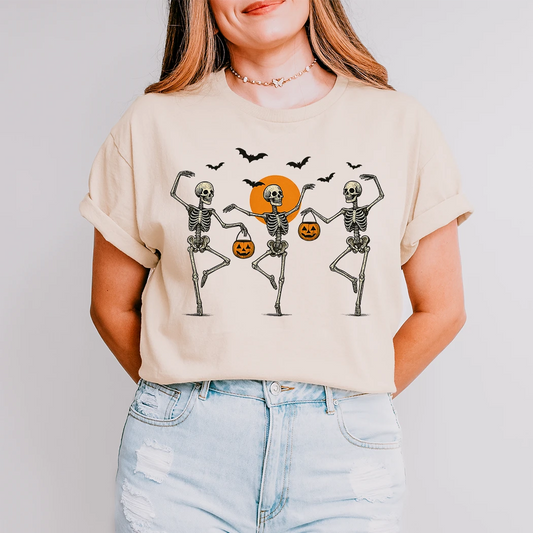 A person stands with their hands behind their back, wearing a cream-colored t-shirt. The shirt has a graphic of three dancing skeletons holding jack-o'-lantern buckets, with a large orange sun and black bats in the background.