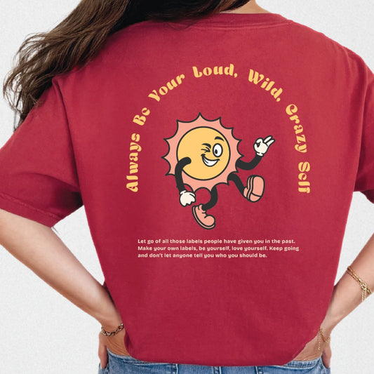Close-up of the back of a maroon t-shirt. A cartoon sun with arms and legs is winking and gesturing with its hands. Arced text above the sun reads 'Always Be Your Loud, Wild, Crazy Self'. Below the sun is a paragraph of smaller white text about being yourself.