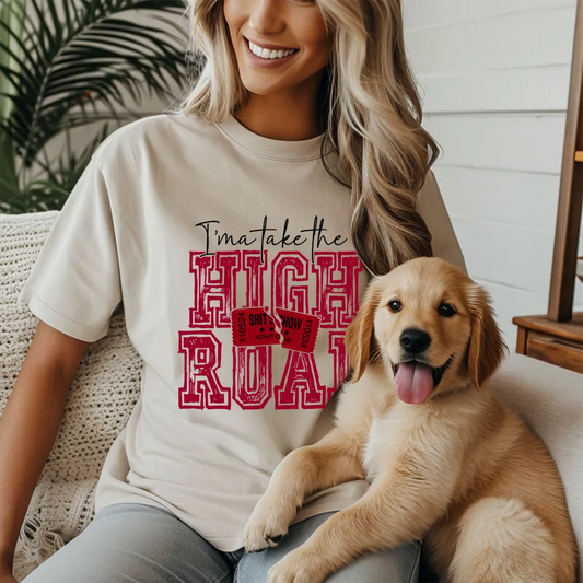A smiling woman with long blonde wavy hair sits on a light-colored textured couch, cuddling a golden retriever puppy. She is wearing a light beige t-shirt with a red graphic that reads "I'ma take the HIGH ROAD" in a stylized font, incorporating red ticket stubs into the design. The puppy is looking directly at the camera with its tongue slightly out. A blurred green leafy plant is visible behind the woman on the left, and a hint of a wooden table or furniture is on the right.
