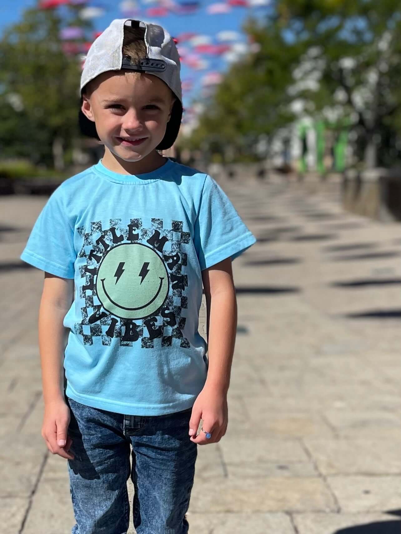 Kid model wearing a blue shirt that says Little Man vibes. The design has a checkered background with yellow smiley face with lightning bulb eyes. 