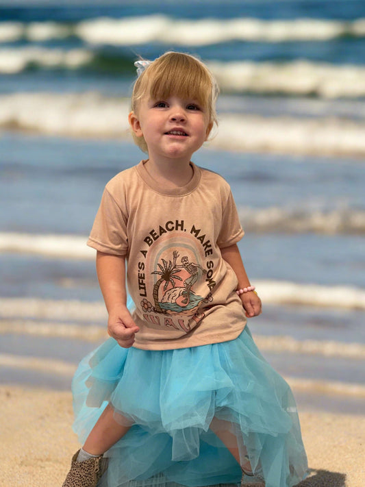 Model kid Tan shirt with design that says Life's A  Beach. Make some waves. It has a skeleton riding a wave with a palm tree. 