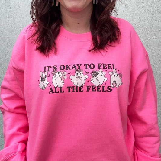 A woman stands against a light-colored wall, smiling and looking to the side. She is wearing a bright, neon pink crewneck with the phrase "IT'S OKAY TO FEEL ALL THE FEELS" and a line of small, multi-colored characters across the chest. She has her hand in the pocket of her light blue denim shorts.







