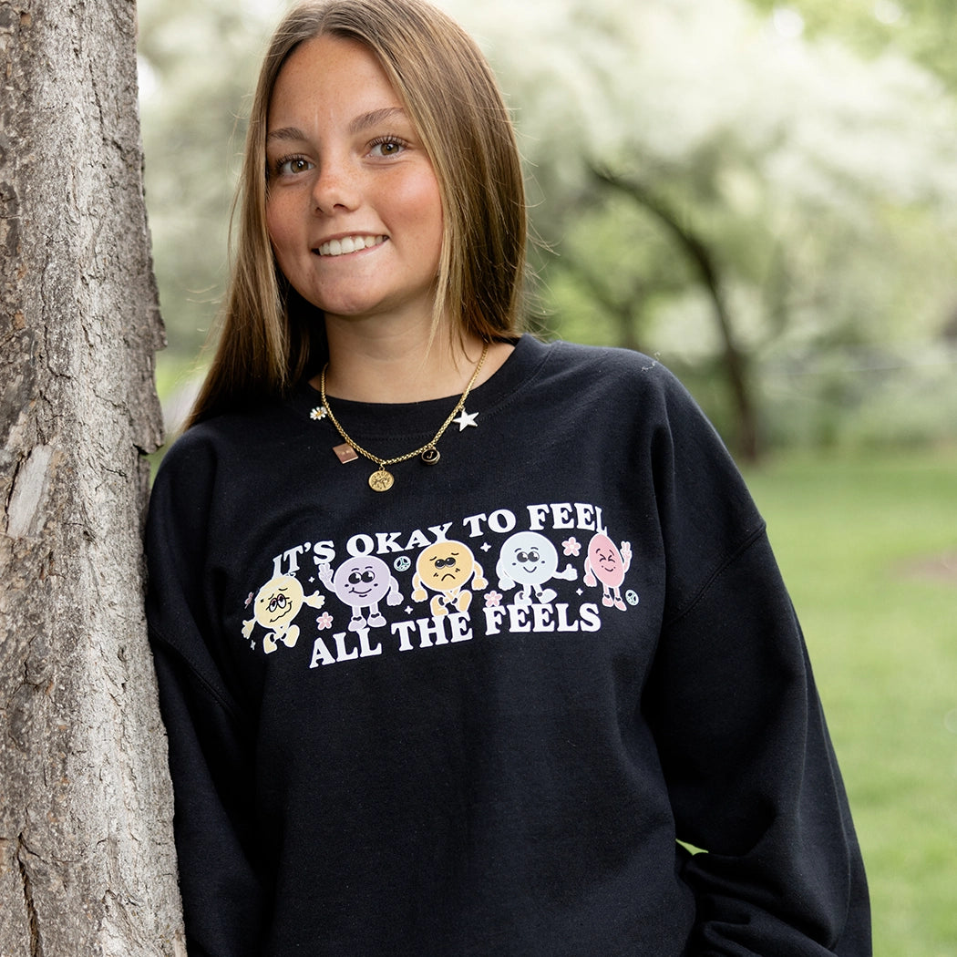 It’s okay to feel all the feels positive message shirt with smiley face characters. Black crewneck with colorful design. 