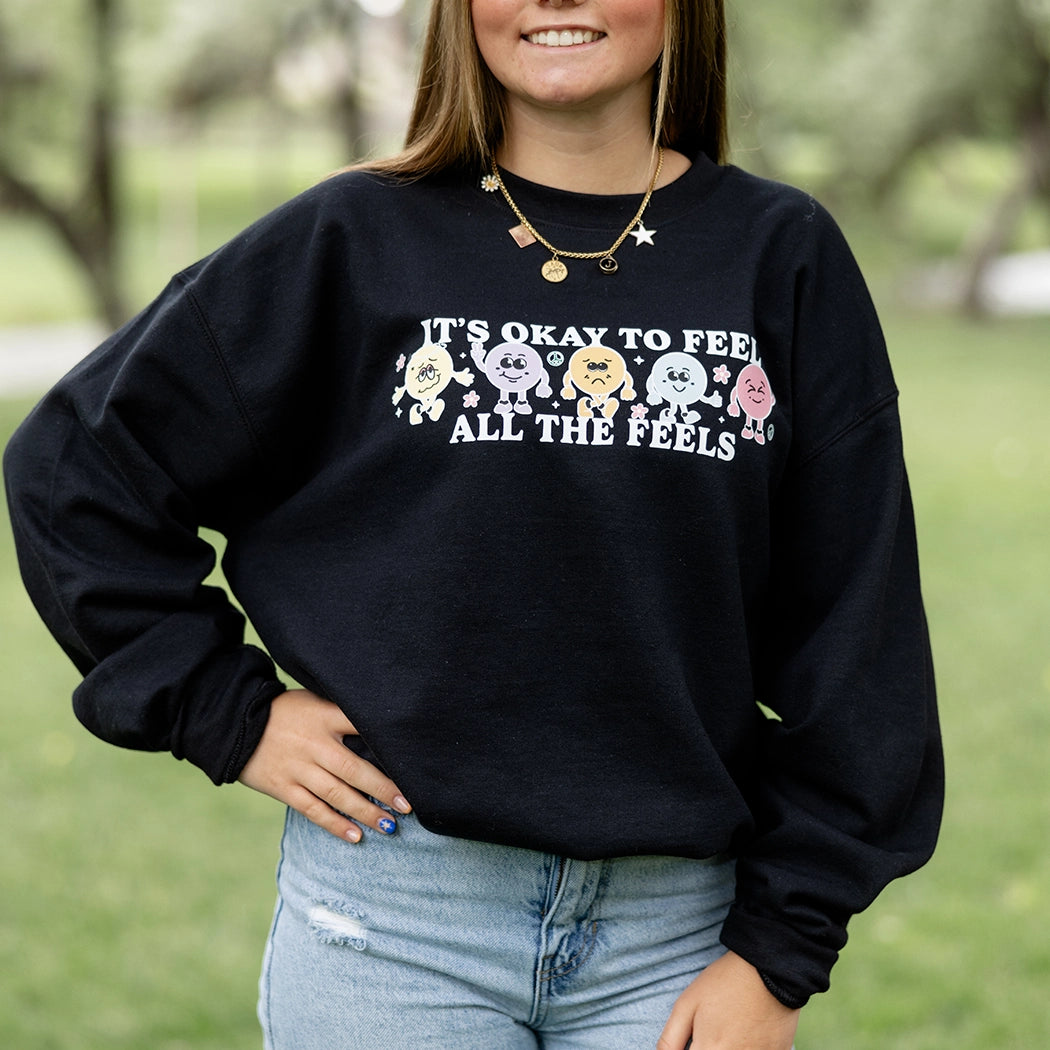 It’s okay to feel all the feels positive message shirt with smiley face characters. Black crewneck with colorful design. 