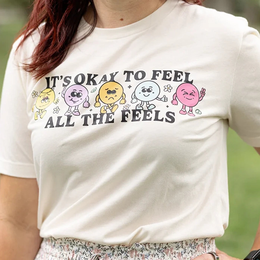 It’s okay to feel all the feels positive message shirt with smiley face characters. White shirt with colorful design. Flowers, peace sign, green, yellow, blue and orange design. 