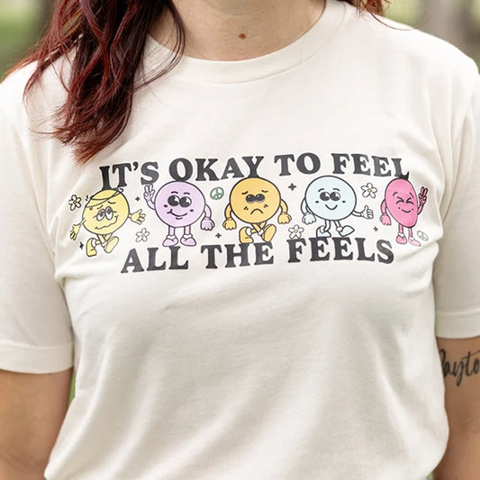 It’s okay to feel all the feels positive message shirt with smiley face characters. White shirt with colorful design. Flowers, peace sign, green, yellow, blue and orange design. 