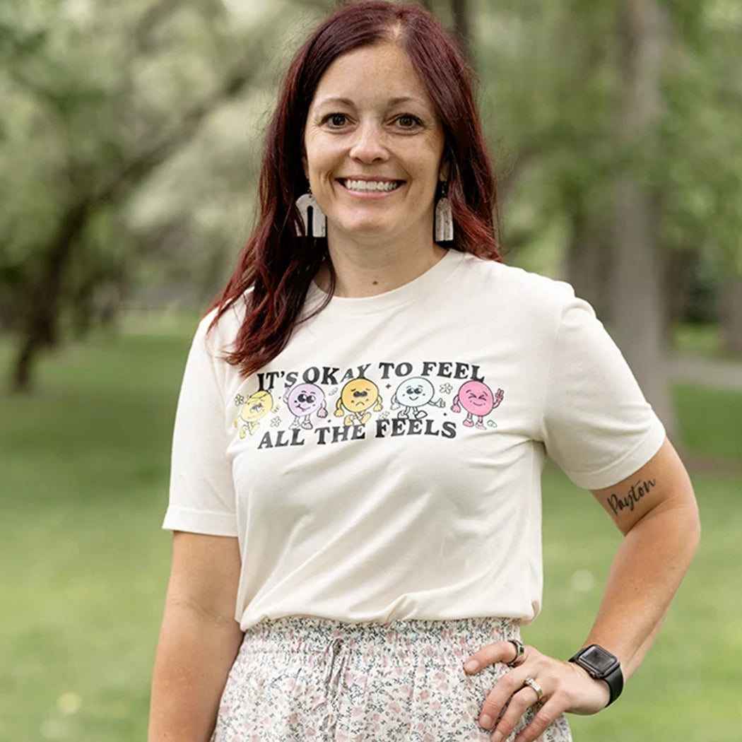 It’s okay to feel all the feels positive message shirt with smiley face characters. White shirt with colorful design. Flowers, peace sign, green, yellow, blue and orange design. 