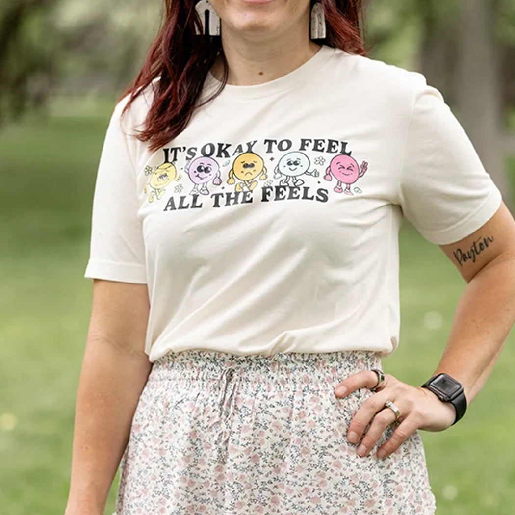 It’s okay to feel all the feels positive message shirt with smiley face characters. White shirt with colorful design. Flowers, peace sign, green, yellow, blue and orange design. 