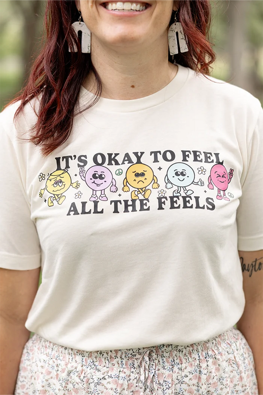 It’s okay to feel all the feels positive message shirt with smiley face characters. White shirt with colorful design. Flowers, peace sign, green, yellow, blue and orange design. 