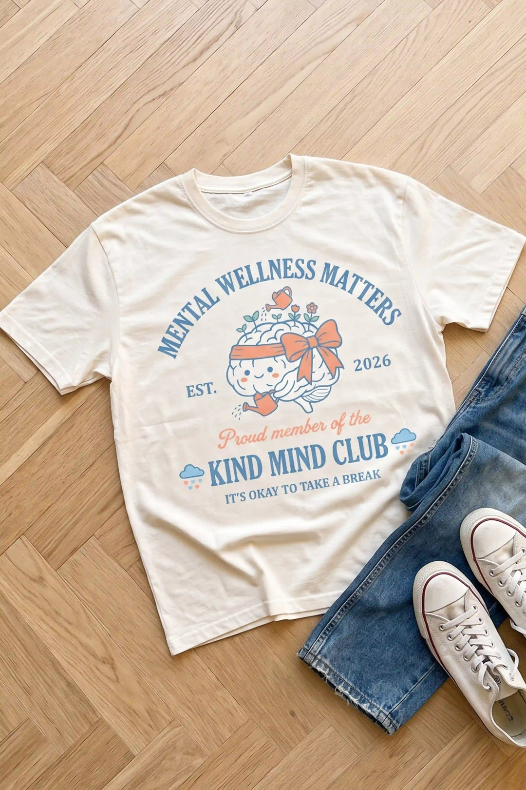 Cream short-sleeve t-shirt laid flat featuring blue arched text reading "MENTAL WELLNESS MATTERS" above a cute brain character wearing a bow and watering flowers, with pink script reading "Proud member of the KIND MIND CLUB"