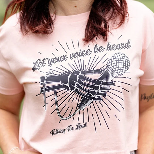 Pink crop top shirt with positive message that says Let Your Voice Be Heard. The design has a Skelton hand with a microphone.  