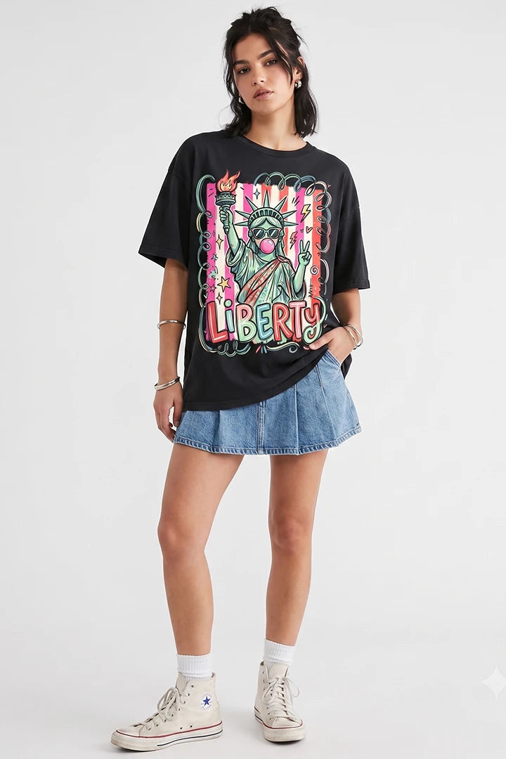 A young woman with dark hair stands against a white background wearing a black, oversized short-sleeved t-shirt. The shirt features a vibrant, colorful cartoon graphic of the Statue of Liberty wearing sunglasses, blowing pink bubblegum, and throwing up a peace sign over a pink-striped background. The word 'LIBERTY' is printed below in colorful bubble letters. She is styled with a blue denim pleated skirt, white socks, and white high-top canvas sneakers.