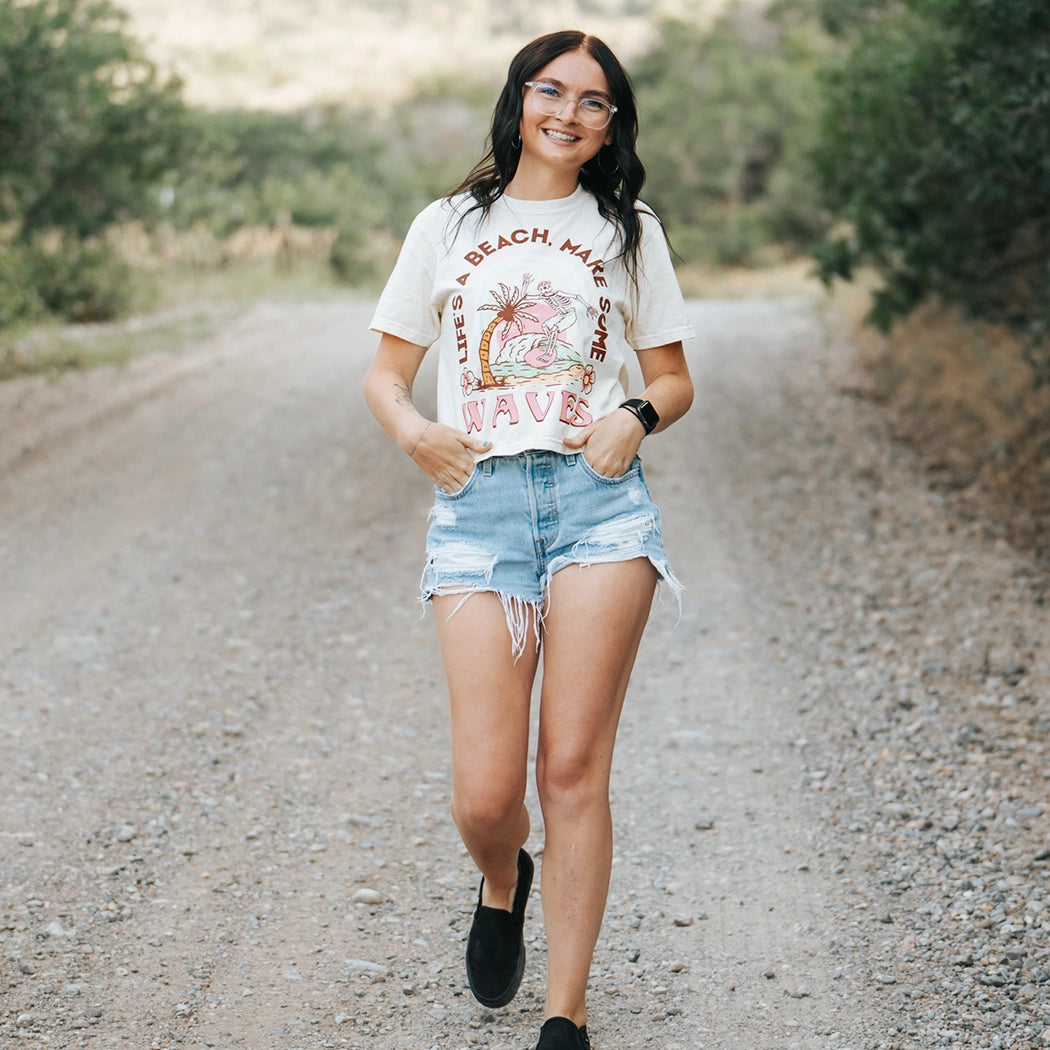 Tan crop top with design that says Life's A  Beach. Make some waves. It has a skeleton riding a wave with a palm tree. 