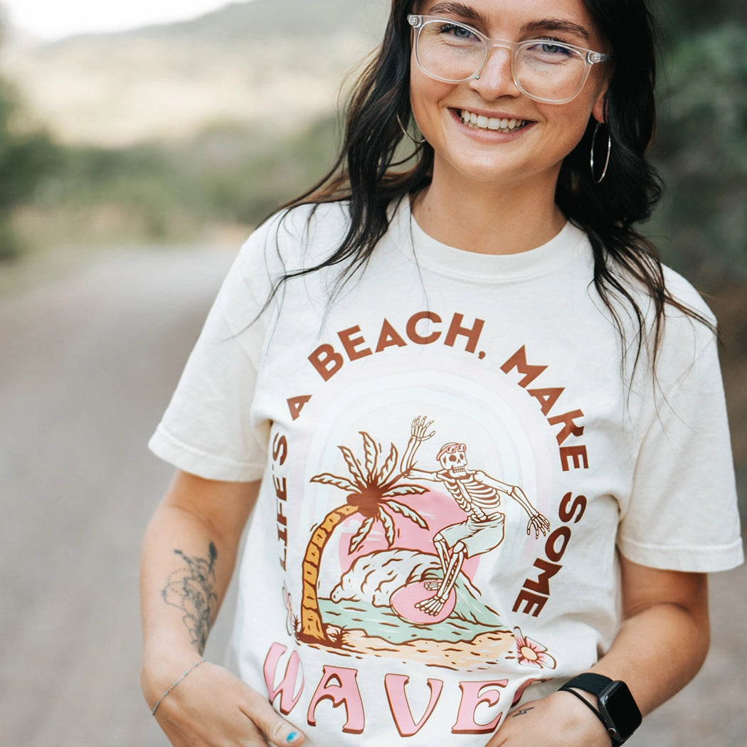 Tan crop top with design that says Life's A  Beach. Make some waves. It has a skeleton riding a wave with a palm tree. 