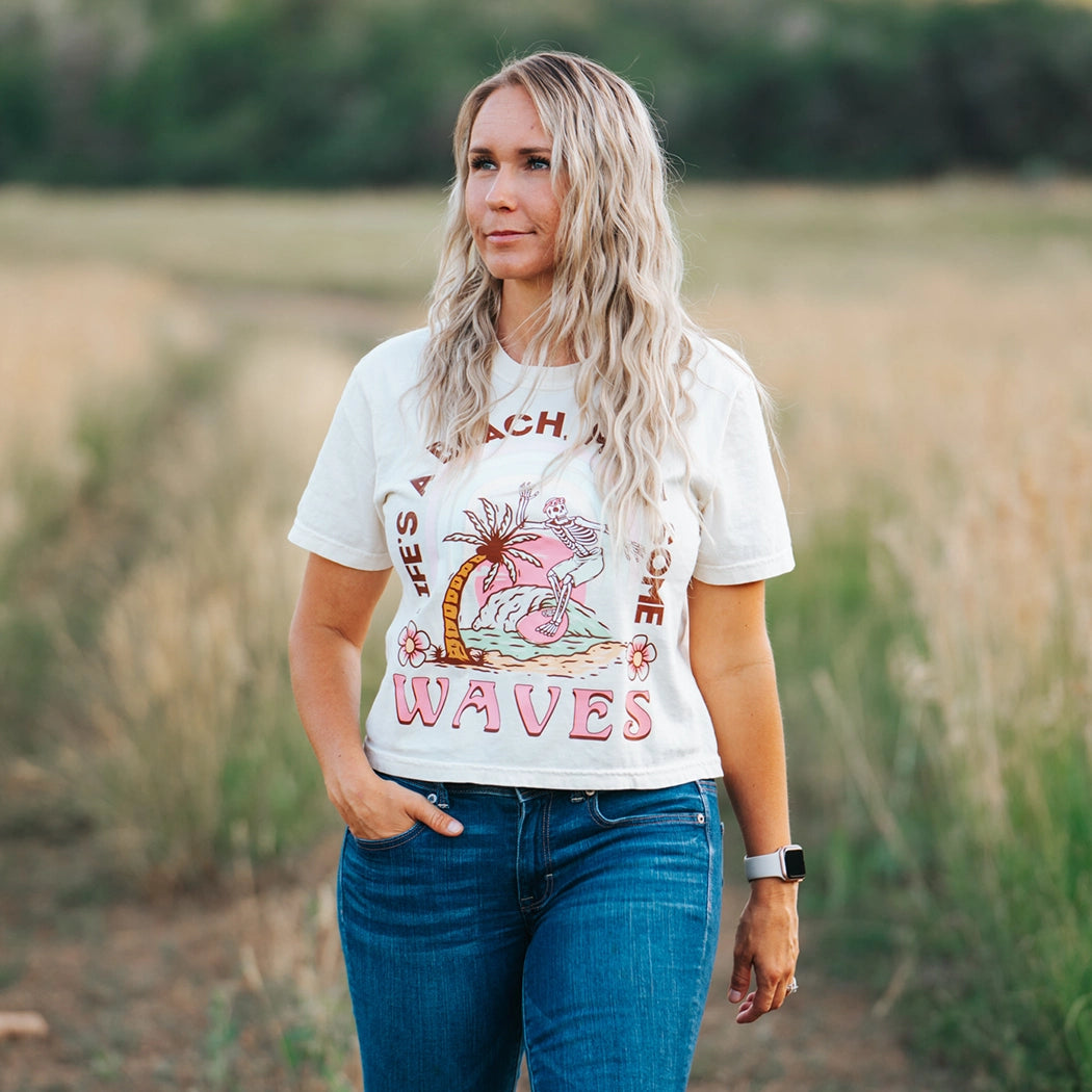 Tan crop top with design that says Life's A  Beach. Make some waves. It has a skeleton riding a wave with a palm tree. 