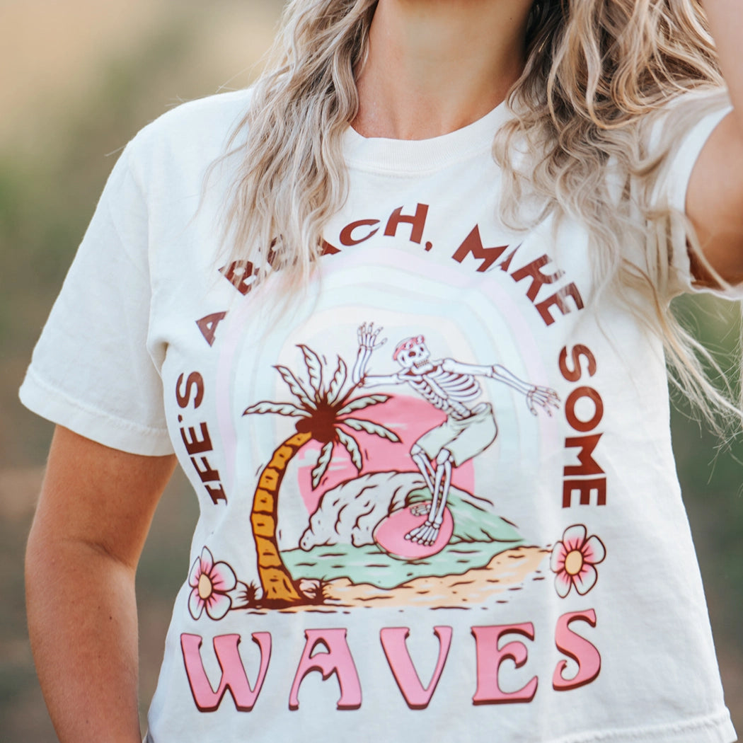 Tan crop top with design that says Life's A  Beach. Make some waves. It has a skeleton riding a wave with a palm tree. 