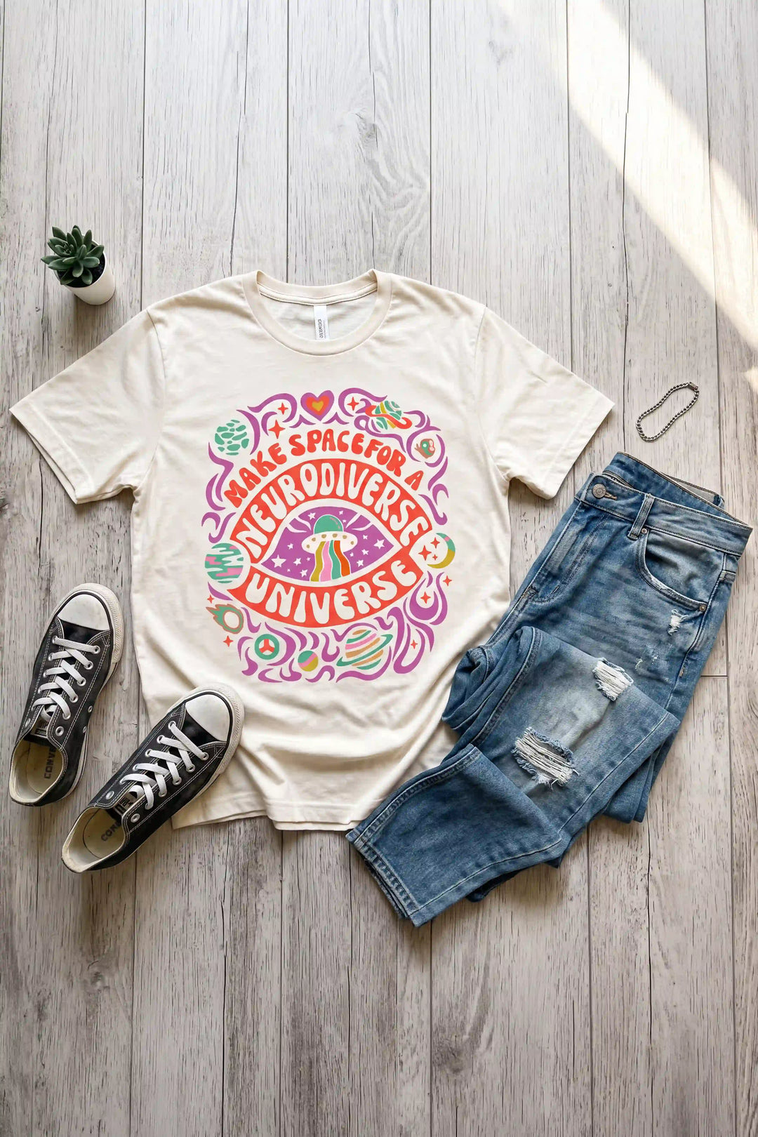 Cream short-sleeve t-shirt laid flat featuring groovy retro 1970s style space art with a flying saucer, planets, stars, and colorful text reading "MAKE SPACE FOR A NEURODIVERSE UNIVERSE"