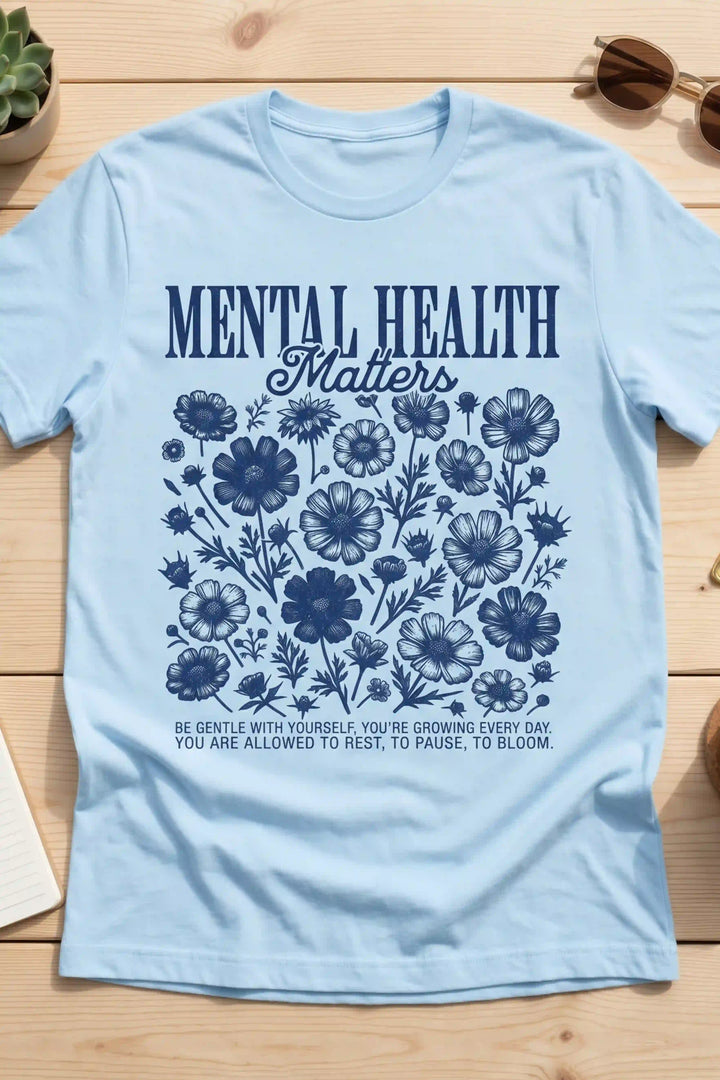 Light blue short-sleeve t-shirt laid flat featuring dark blue text reading "MENTAL HEALTH Matters" above detailed botanical wildflower line art and self-care text