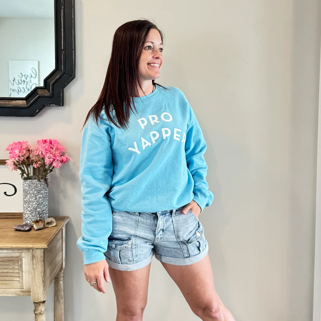 A fair-skinned person with shoulder-length dark hair smiles to their right, wearing a light blue crewneck sweatshirt with the words "PRO" stacked above "YAPPER" in a simple, rounded white font on the chest. graphic tee hoodies